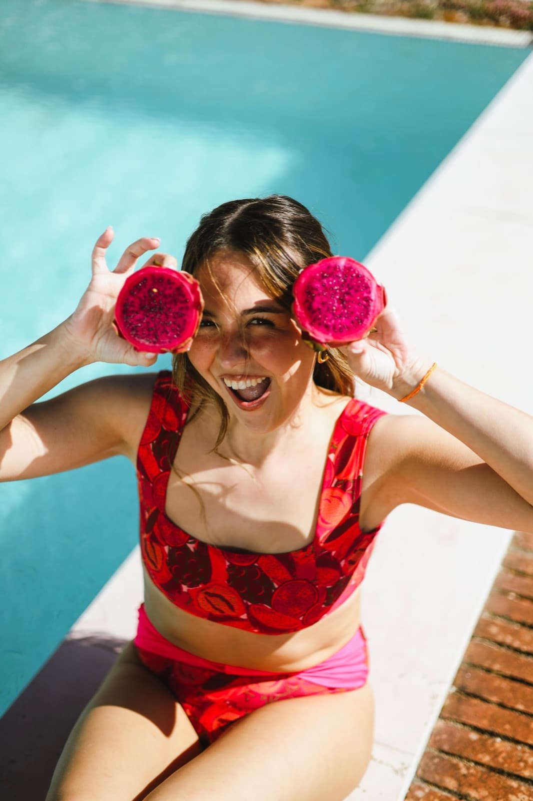 Mulher à beira da piscina a rir, com duas metades de pitaya à frente dos olhos, com o biquíni Oceana em Pitaya